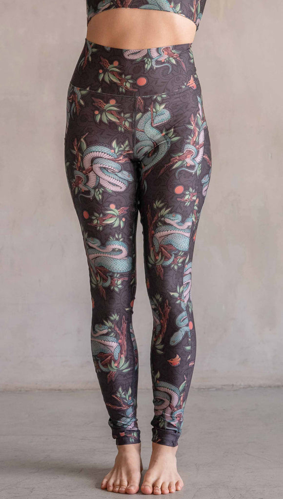 Model wearing WERKSHOP Pit Viper Featherlight Leggings. The artwork on the leggings have clusters of teal blue pit viper snakes intertwined on tree branches over a taupe/brown background. The featherlight leggings have pockets.