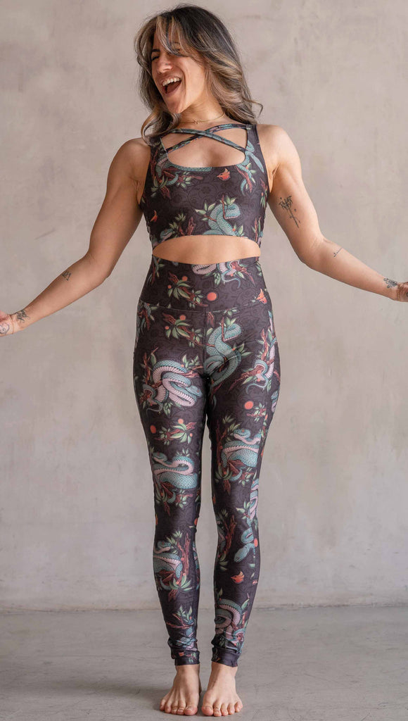 Model wearing WERKSHOP Pit Viper Featherlight Leggings. The artwork on the leggings have clusters of teal blue pit viper snakes intertwined on tree branches over a taupe/brown background. The featherlight leggings have pockets.