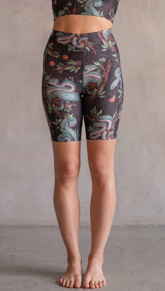 Model wearing WERKSHOP Pit Viper Featherlight Bicycle Length shorts. The artwork on the shorts have clusters of teal blue pit viper snakes intertwined on tree branches over a taupe/brown background. The featherlight shorts have pockets.