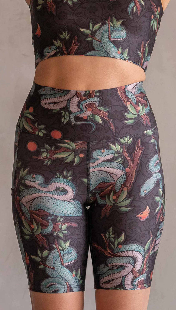 Model wearing WERKSHOP Pit Viper Featherlight Bicycle Length shorts. The artwork on the shorts have clusters of teal blue pit viper snakes intertwined on tree branches over a taupe/brown background. The featherlight shorts have pockets.