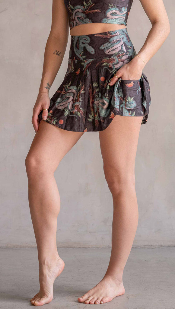Model wearing WERKSHOP Pit Viper Featherlight Active Skirt. The skirts have built in shorts underneath and artwork on the shorts have clusters of teal blue pit viper snakes intertwined on tree branches over a taupe/brown background. The featherlight skirts have pockets.