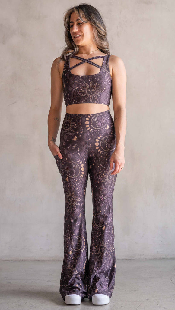 Model wearing WERKSHOP Zodiac Bell Bottoms. They are high waist and feature pockets on both legs. The zodiac themed artwork shows a hand-drawn sun and moon with the moon phases, shooting stars and all 12 zodiac constellations in gold over a dark purple background. Our model is 5’2” and this images shows her wearing the bells with sneakers. The hem of the pants are hitting the floor.