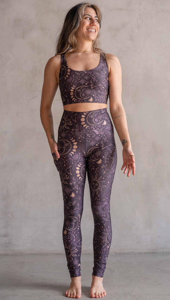 Model wearing WERKSHOP Zodiac Leggings. They are high waist and feature pockets on both legs. The zodiac themed artwork shows a hand-drawn sun and moon with the moon phases, shooting stars and all 12 zodiac constellations in gold over a dark purple background.