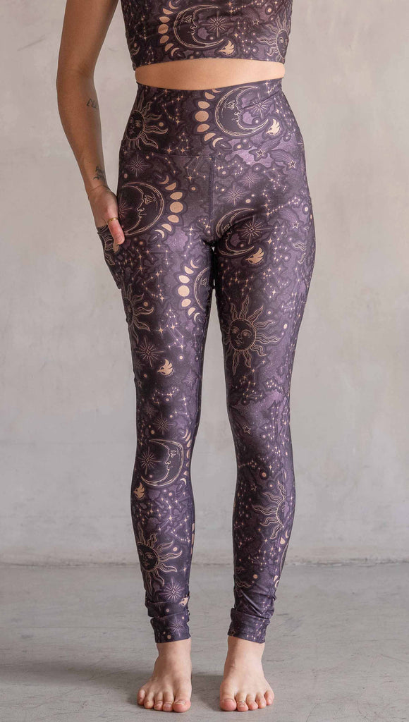 Model wearing WERKSHOP Zodiac Leggings. They are high waist and feature pockets on both legs. The zodiac themed artwork shows a hand-drawn sun and moon with the moon phases, shooting stars and all 12 zodiac constellations in gold over a dark purple background.