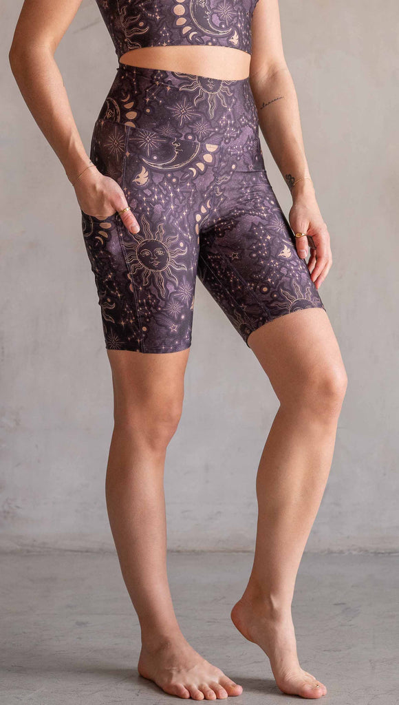 Model wearing WERKSHOP Zodiac Bicycle Length Shorts. They are high waist and feature pockets on both legs. The zodiac themed artwork shows a hand-drawn sun and moon with the moon phases, shooting stars and all 12 zodiac constellations in gold over a dark purple background. The length of the shorts hit in the mid thigh, above the knee.