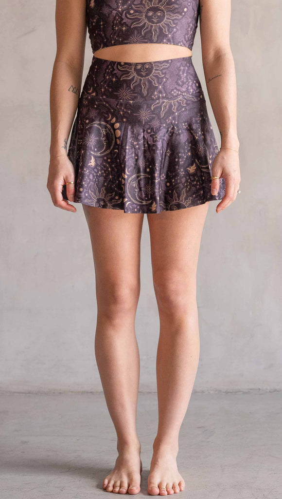 Model wearing WERKSHOP Zodiac Tennis Skirt with built-in shorts. They are high waist and feature pockets on both legs under the flirty skirt. The zodiac themed artwork shows a hand-drawn sun and moon with the moon phases, shooting stars and all 12 zodiac constellations in gold over a dark purple background. The skirt length hits right around the models fingertips.