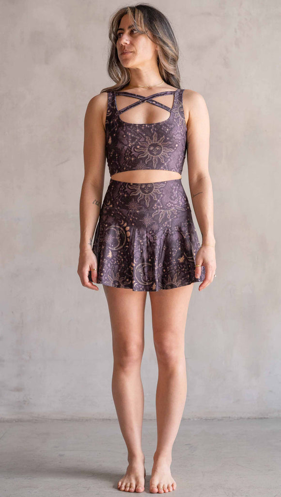Model wearing WERKSHOP Zodiac Tennis Skirt with built-in shorts. They are high waist and feature pockets on both legs under the flirty skirt. The zodiac themed artwork shows a hand-drawn sun and moon with the moon phases, shooting stars and all 12 zodiac constellations in gold over a dark purple background. The skirt length hits right around the models fingertips.