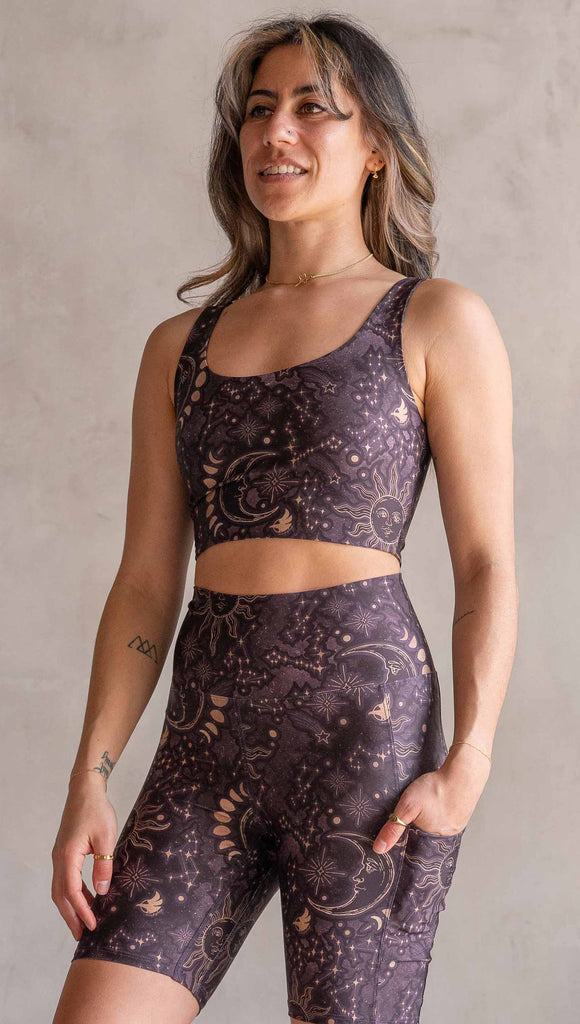 Front view of model wearing WERKSHOP Four-Way Reversible Top. The top has a tarot card themed artwork on a dark blue background on one side and a zodiac themed artwork on purple background on the opposite side. She is wearing it with the Purple side out and the X neckline detail in the back.
