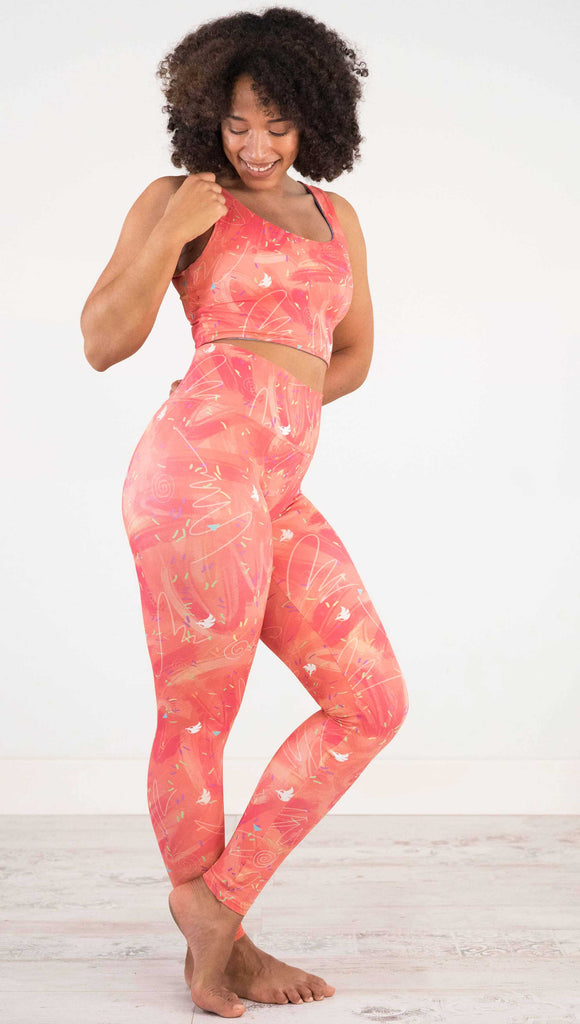 Full body side view of model wearing WERKSHOP Salmon Scribble Leggings with coral  brushstrokes over a bright salmon background. Also has little confetti and eagle logos scattered throughout.