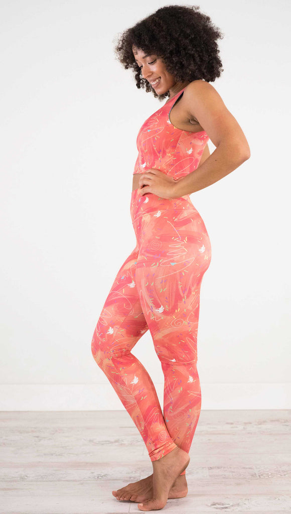 Full body side view of model wearing WERKSHOP Salmon Scribble Leggings with coral  brushstrokes over a bright salmon background. Also has little confetti and eagle logos scattered throughout.