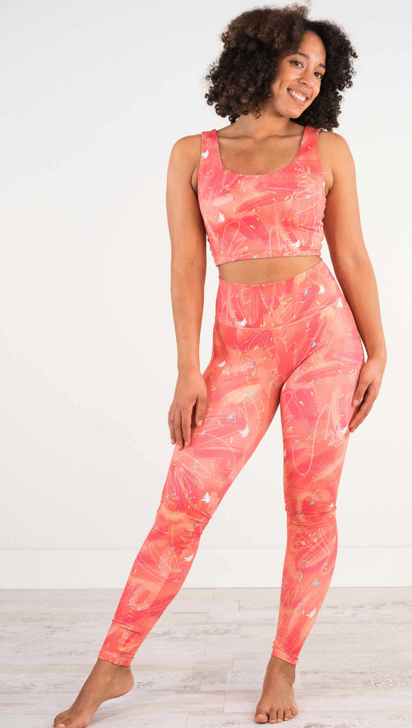 Full body front view of model wearing WERKSHOP Salmon Scribble Leggings with coral  brushstrokes over a bright salmon background. Also has little confetti and eagle logos scattered throughout.