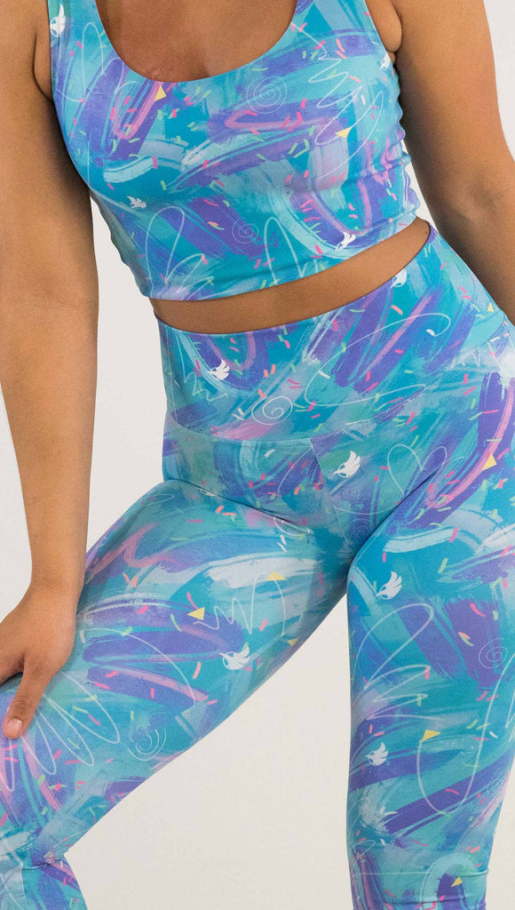 zoomed-in view of model wearing WERKSHOP Teal Scribble Leggings with purple and pink brushstrokes over a bright teal background. Also has little confetti and eagle logos scattered throughout.