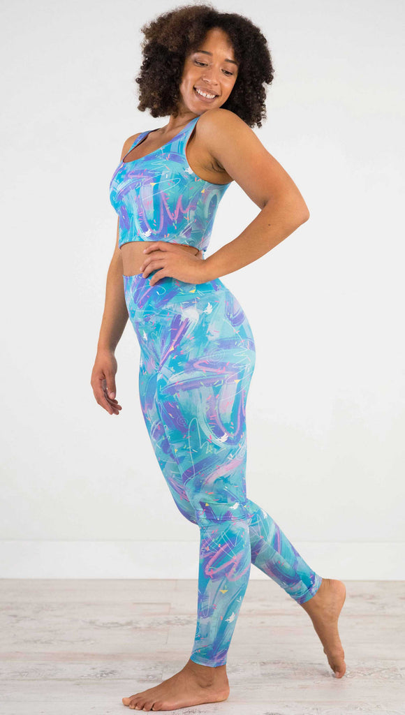 Full body side view of model wearing WERKSHOP Teal Scribble Leggings with purple and pink brushstrokes over a bright teal background. Also has little confetti and eagle logos scattered throughout.