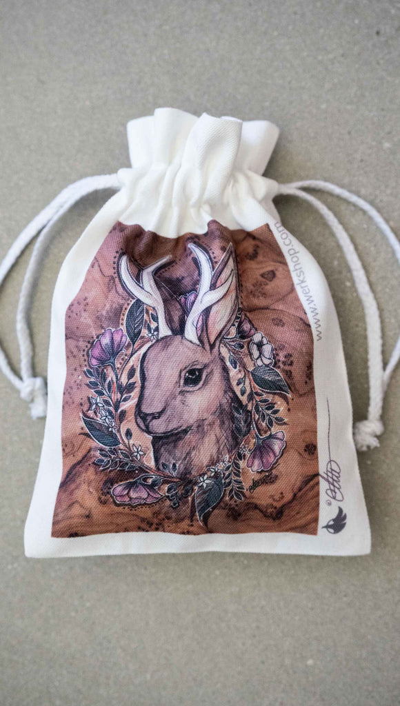Small (Matching) canvas drawstring pouch that comes free with the purchase of a puzzle. The puzzle is printed with original artwork of a Jackalope by Chriztina Marie. The Jackalope is a mythological creature that looks like a bunny with deer antlers. The illustration shows the head of a Jackalope over a wood grain background with a wreathe of flowers. 