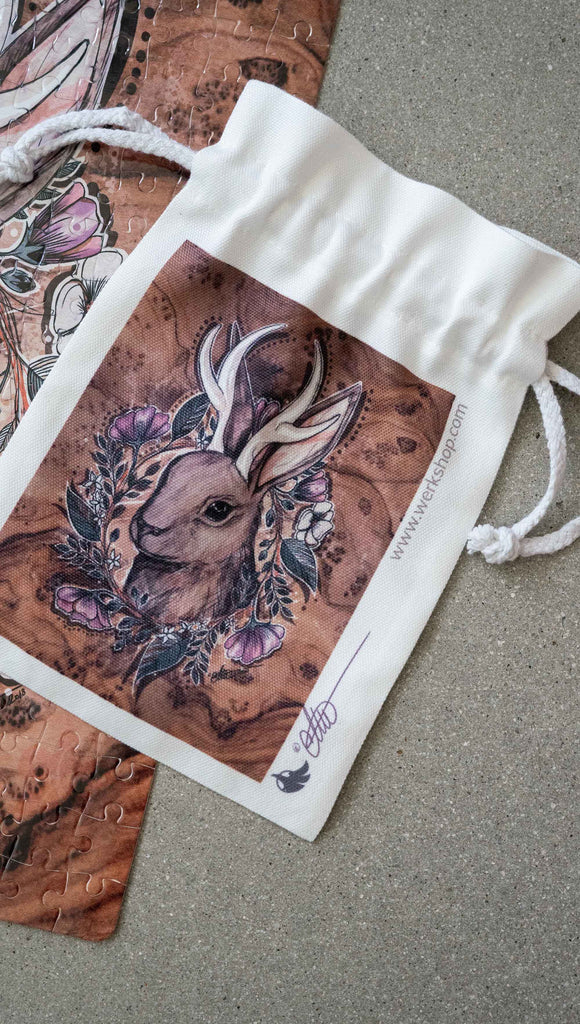 Small (Matching) canvas drawstring pouch that comes free with the purchase of a puzzle. The puzzle is printed with original artwork of a Jackalope by Chriztina Marie. The Jackalope is a mythological creature that looks like a bunny with deer antlers. The illustration shows the head of a Jackalope over a wood grain background with a wreathe of flowers.