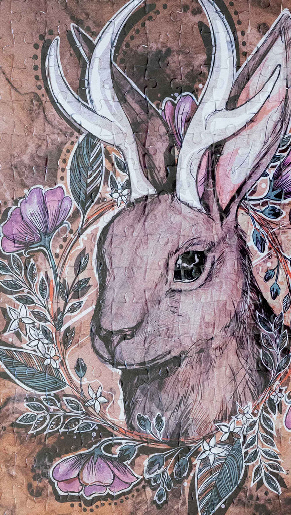 Zoomed in image of WERKSHOP Jackalope 252 piece jigsaw puzzle. The puzzle is printed with original artwork of a Jackalope by Chriztina Marie. The Jackalope is a mythological creature that looks like a bunny with deer antlers. The illustration shows the head of a Jackalope over a wood grain background with a wreathe of flowers. 