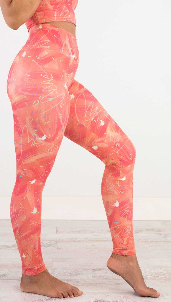 Waist down side view of model wearing WERKSHOP Salmon Scribble Leggings with coral  brushstrokes over a bright salmon background. Also has little confetti and eagle logos scattered throughout.