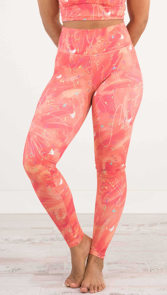 Waist down front view of model wearing WERKSHOP Salmon Scribble Leggings with coral  brushstrokes over a bright salmon background. Also has little confetti and eagle logos scattered throughout.