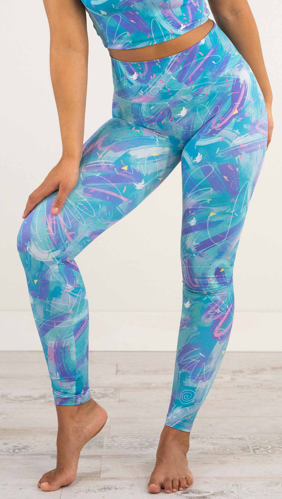 Waist down front view of model wearing WERKSHOP Teal Scribble Leggings with purple and pink brushstrokes over a bright teal background. Also has little confetti and eagle logos scattered throughout.
