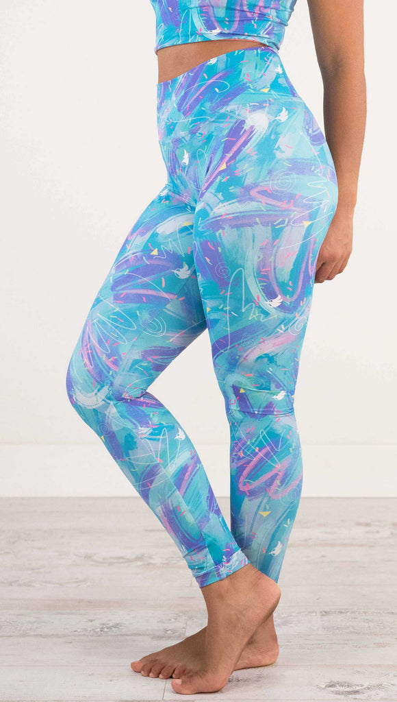 Waist down side view of model wearing WERKSHOP Teal Scribble Leggings with purple and pink brushstrokes over a bright teal background. Also has little confetti and eagle logos scattered throughout.