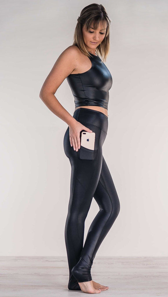 Side view of model wearing shiny black leggings putting iphone into right side pocket