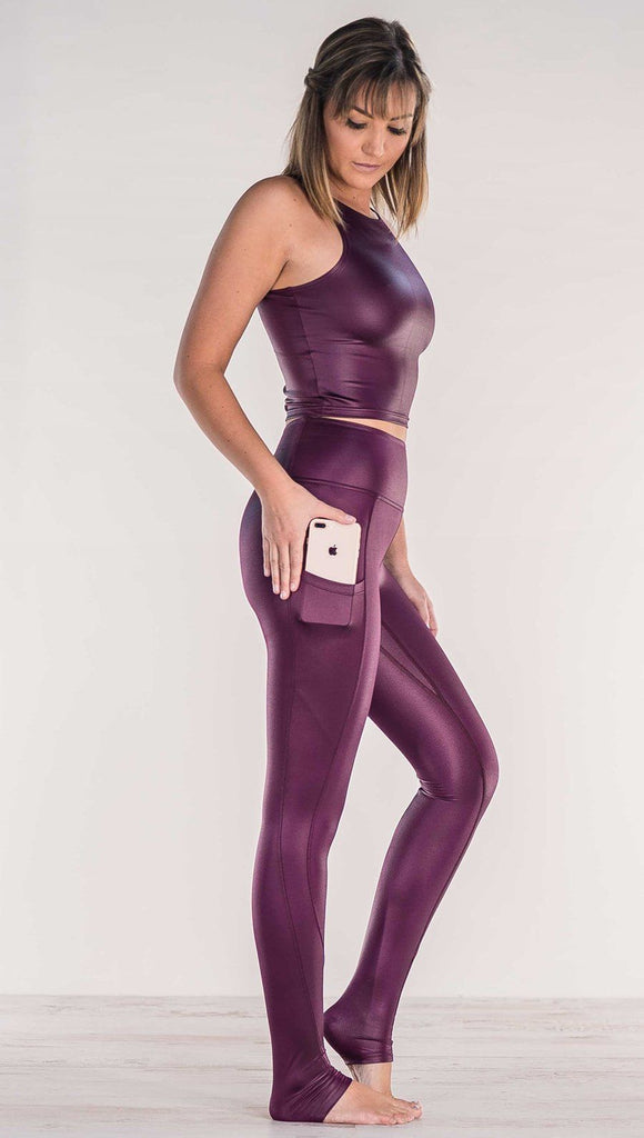 Side view of model wearing shiny eggplant purple colored full length leggings putting iphone into right side pocket