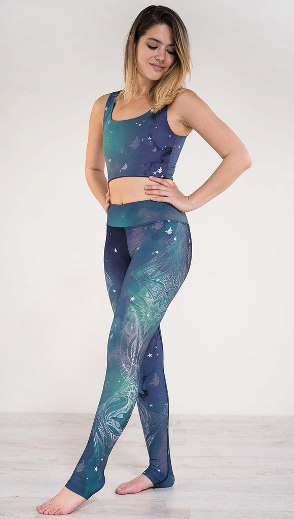 Model pointing toe wearing a blue and green galaxy themed triathlon leggings with white henna inspired art running along the left side of the leg