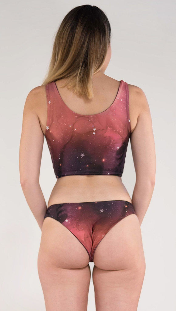 Back side view of model wearing reversible low rise red, orange and purple galaxy themed bikini bottom called Galactic Solar Flare on this side