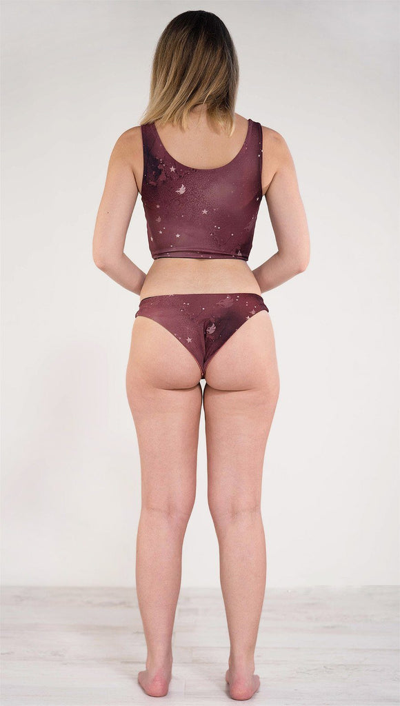 Back side view of model wearing reversible low rise merlot color galaxy themed bikini bottom called Galactic Merlot on this side