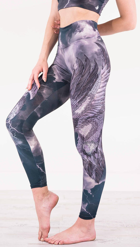Left view of model wearing dark gray athleisure leggings with a large purple bird across the leg with lightning in the background