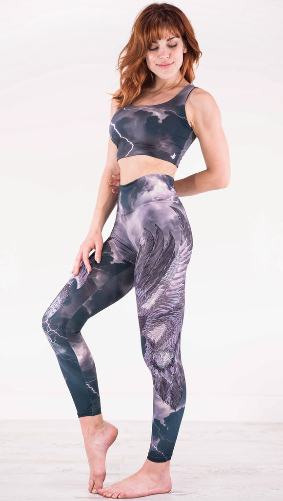 Left view of model wearing dark gray athleisure leggings with a large purple bird across the leg with lightning in the background