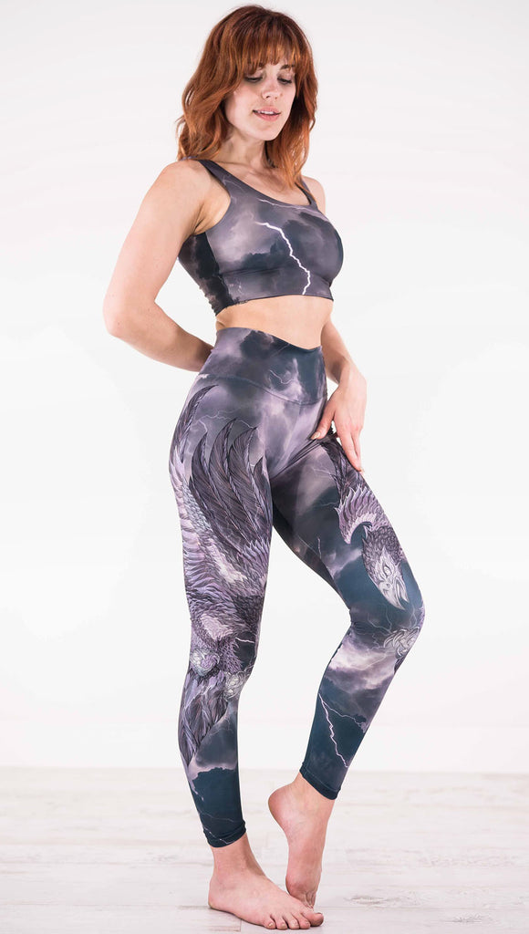 Right view of model wearing dark gray athleisure leggings with a large purple bird across the leg with lightning in the background