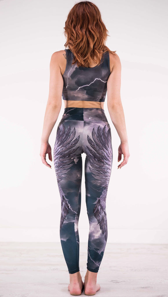 Back view of model wearing dark gray athleisure leggings with a large purple bird across each leg with lightning in the background
