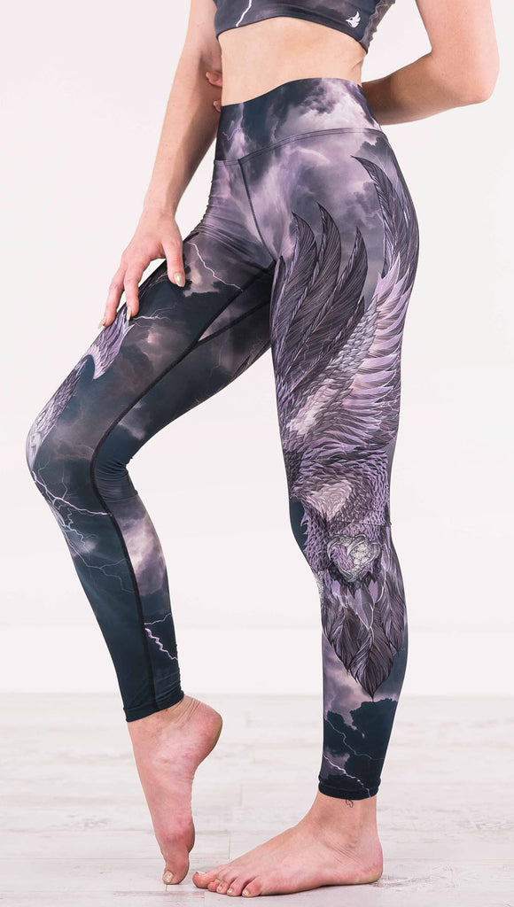 Left view of model wearing dark gray triathlon leggings with a large purple bird across the leg with lightning in the background