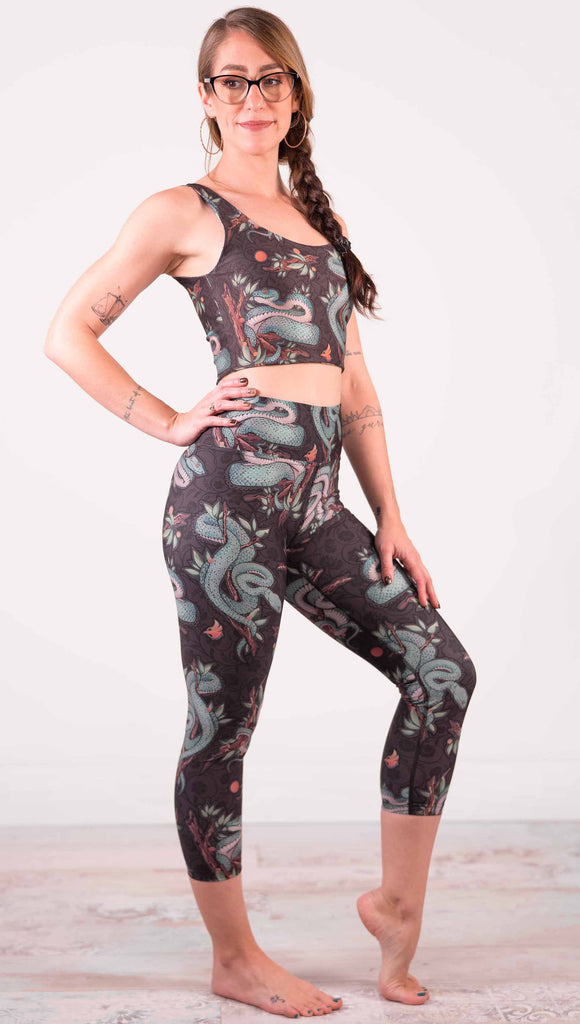 Model wearing WERKSHOP Pit Viper Triathlon Leggings. The artwork on the leggings have clusters of teal blue pit viper snakes intertwined on tree branches over a taupe/brown background