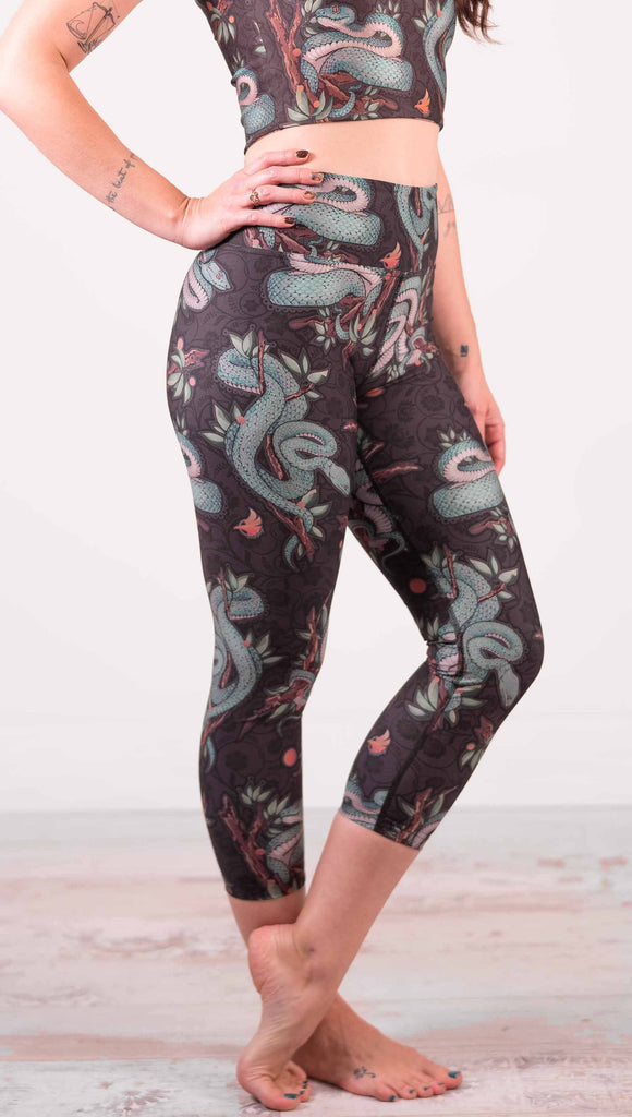 Model wearing WERKSHOP Pit Viper Triathlon Leggings. The artwork on the leggings have clusters of teal blue pit viper snakes intertwined on tree branches over a taupe/brown background