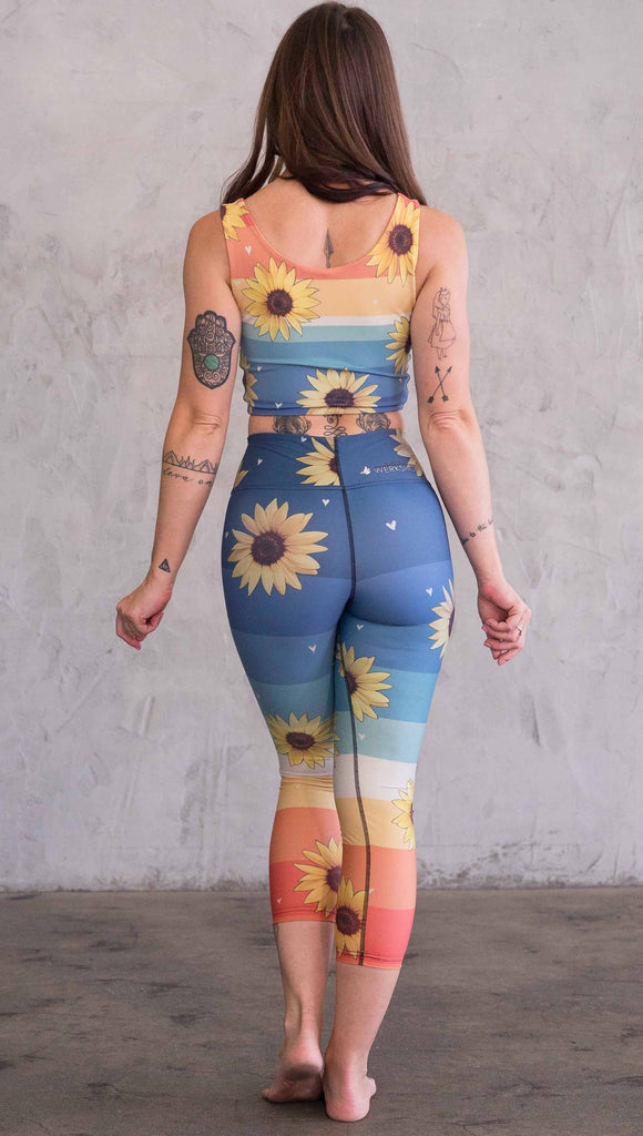 Full body back view of model wearing WERKSHOP Sunflower triathlon capri leggings. The leggings have wide horizontal stripes with dark blue at the waistband, to aqua and pale green at the mid thigh leading to cream at the knee and orange and red tones to the ankle. There are large photo-real sunflowers and tiny hand sketched off-white hearts sprinkled throughout.