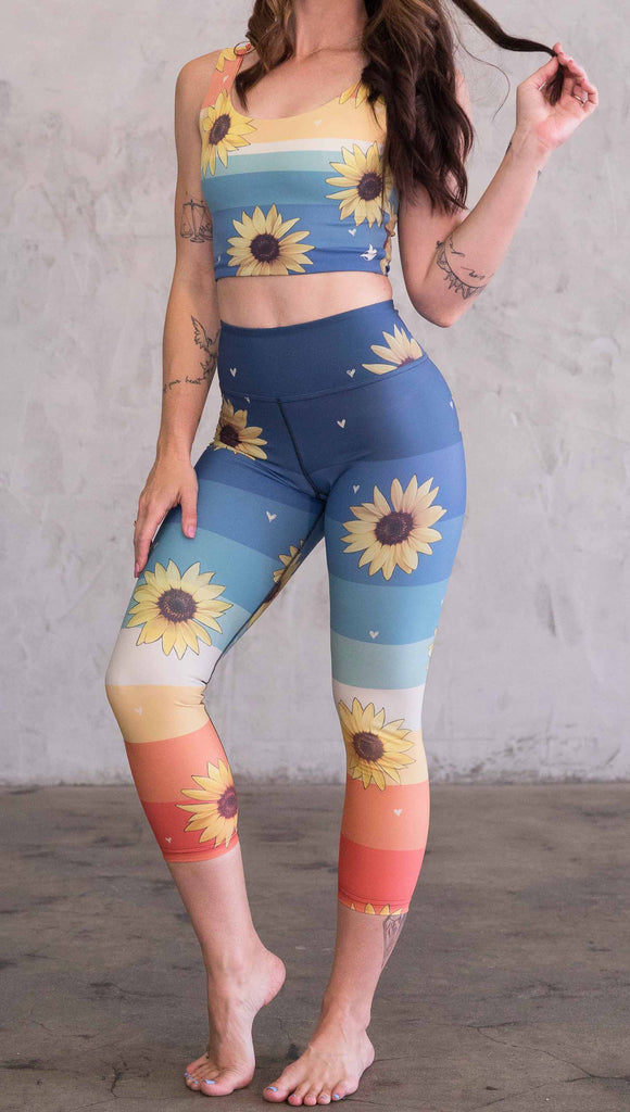 Full body front view of model wearing WERKSHOP Sunflower triathlon capri leggings. The leggings have wide horizontal stripes with dark blue at the waistband, to aqua and pale green at the mid thigh leading to cream at the knee and orange and red tones to the ankle. There are large photo-real sunflowers and tiny hand sketched off-white hearts sprinkled throughout.