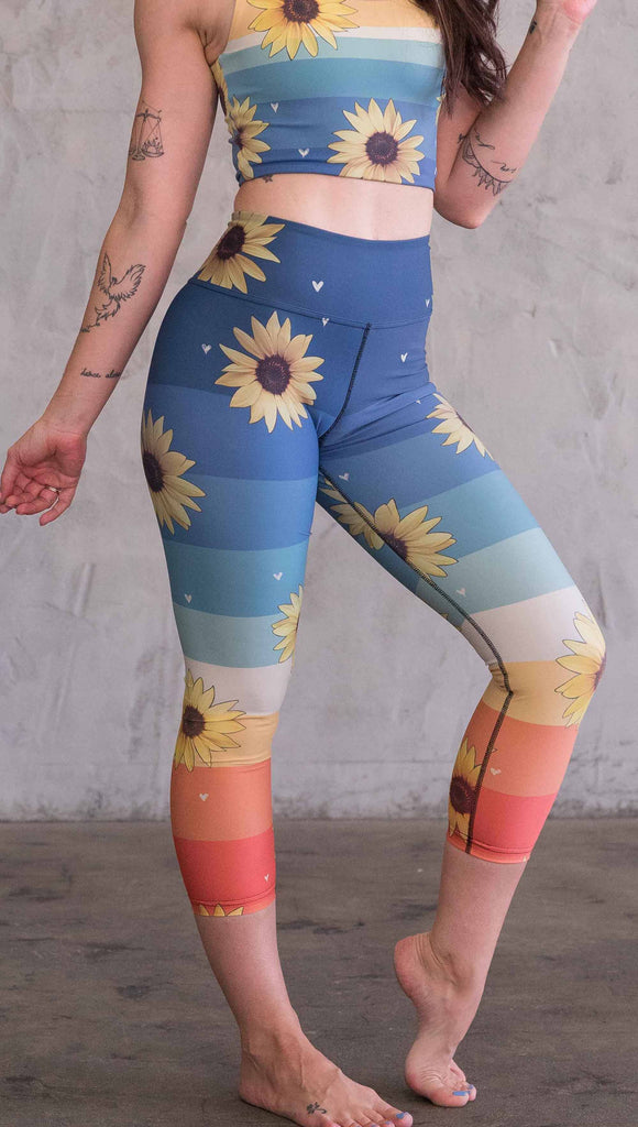 Waist down front view of model wearing WERKSHOP Sunflower triathlon capri leggings. The leggings have wide horizontal stripes with dark blue at the waistband, to aqua and pale green at the mid thigh leading to cream at the knee and orange and red tones to the ankle. There are large photo-real sunflowers and tiny hand sketched off-white hearts sprinkled throughout.