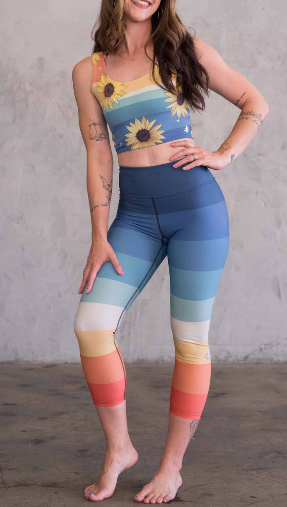 Full body front view of model wearing WERKSHOP Vintage Rainbow Triathlon Capri leggings. The leggings have wide horizontal stripes with dark blue at the waistband, to auqua and pale green at the mid thigh leading to cream at the knee and orange and red tones to the ankle.