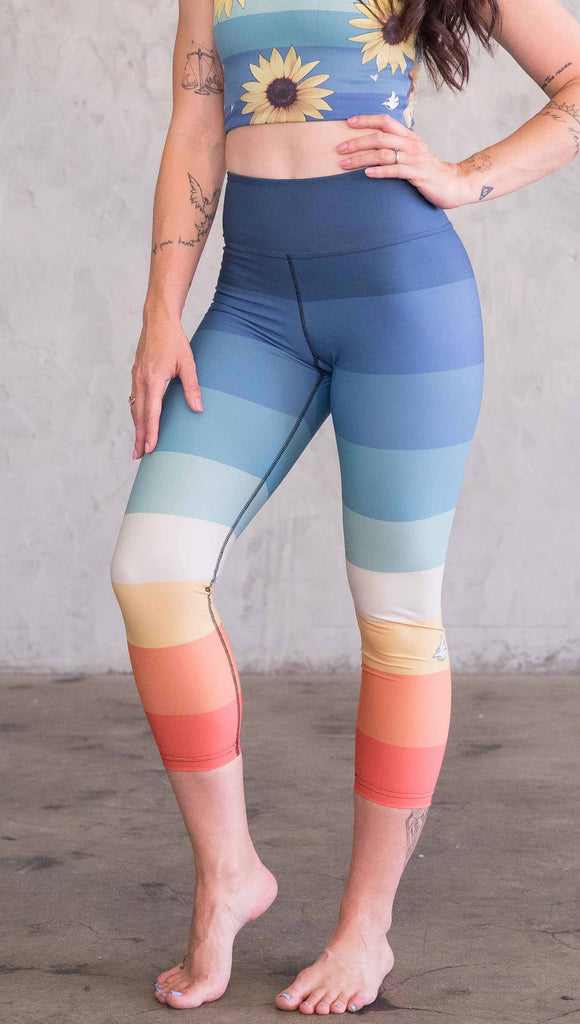 Waist down front view of model wearing WERKSHOP Vintage Rainbow Triathlon Capri leggings. The leggings have wide horizontal stripes with dark blue at the waistband, to auqua and pale green at the mid thigh leading to cream at the knee and orange and red tones to the ankle.