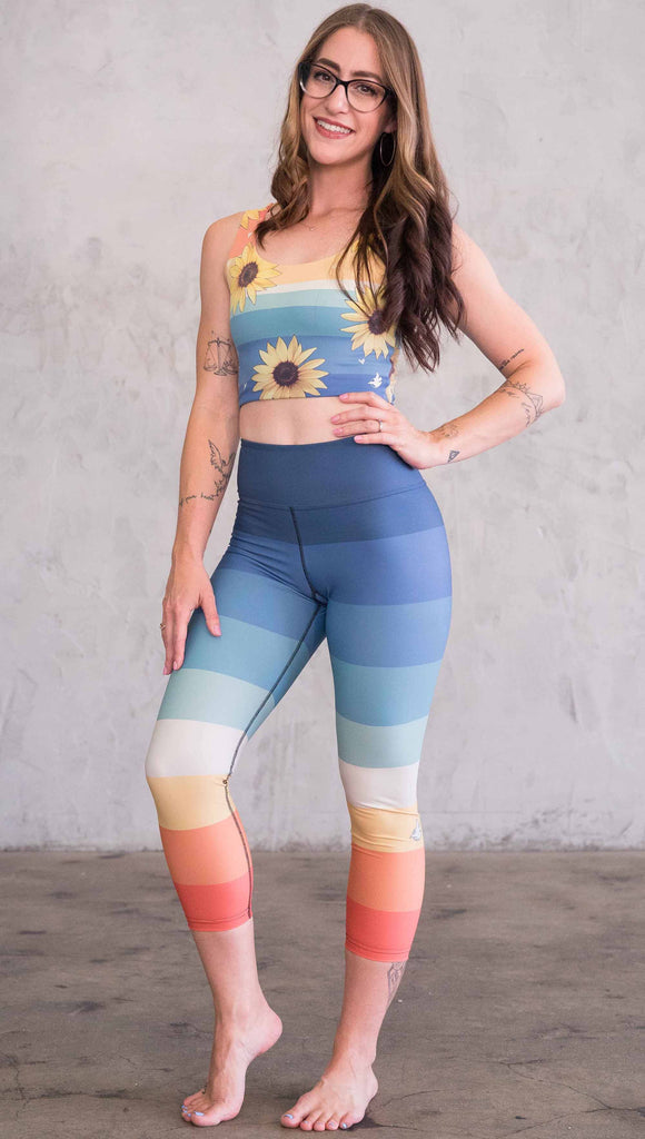 Full body front view of model wearing WERKSHOP Vintage Rainbow Triathlon Capri leggings. The leggings have wide horizontal stripes with dark blue at the waistband, to auqua and pale green at the mid thigh leading to cream at the knee and orange and red tones to the ankle.
