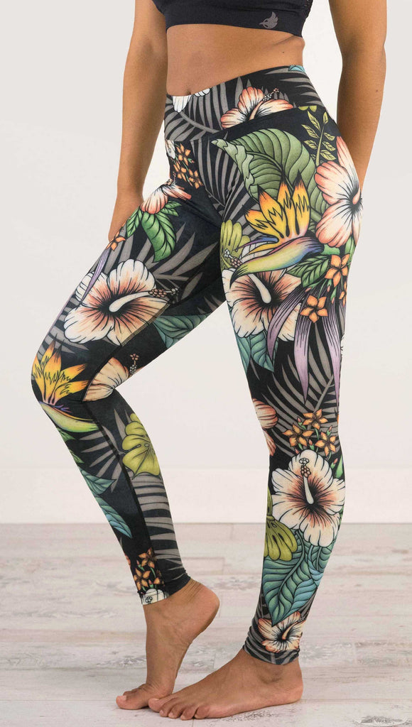 Waist down and side view of model wearing WERKSHOP Floral Night Full Length Triathlon Leggings. The artwork on the leggings has tropical flowers (bird of paradise, hibiscus and palm leaves) on a distressed black background.