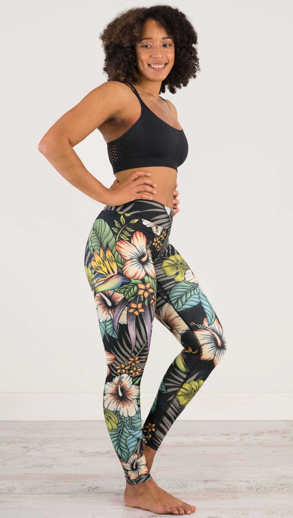 Full body side view of model wearing WERKSHOP Floral Night Full Length Triathlon Leggings. The artwork on the leggings has tropical flowers (bird of paradise, hibiscus and palm leaves) on a distressed black background.