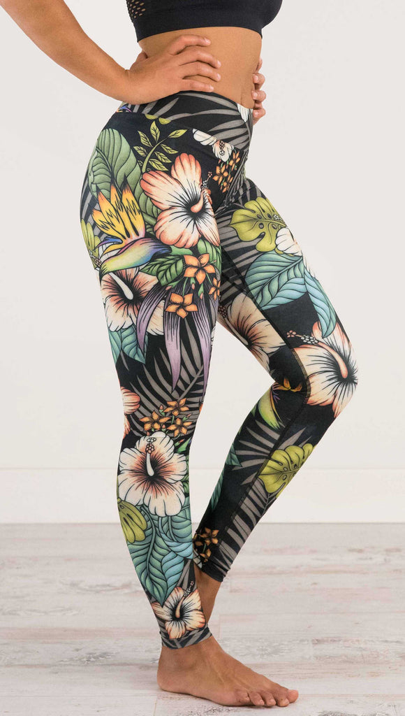 Waist down and side view of model wearing WERKSHOP Floral Night Full Length Triathlon Leggings. The artwork on the leggings has tropical flowers (bird of paradise, hibiscus and palm leaves) on a distressed black background.