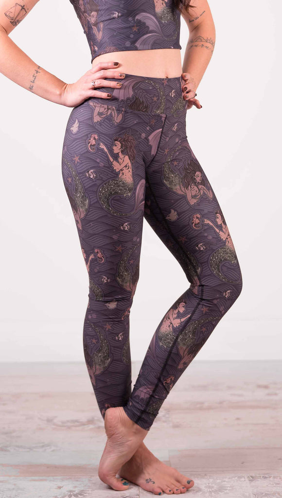 Model wearing WERKSHOP full length triathlon leggings with Original Mermaids artwork. The mermaids have small intricate details on the fins and are swimming with seahorses and angel fish over a dark blue background with waves.