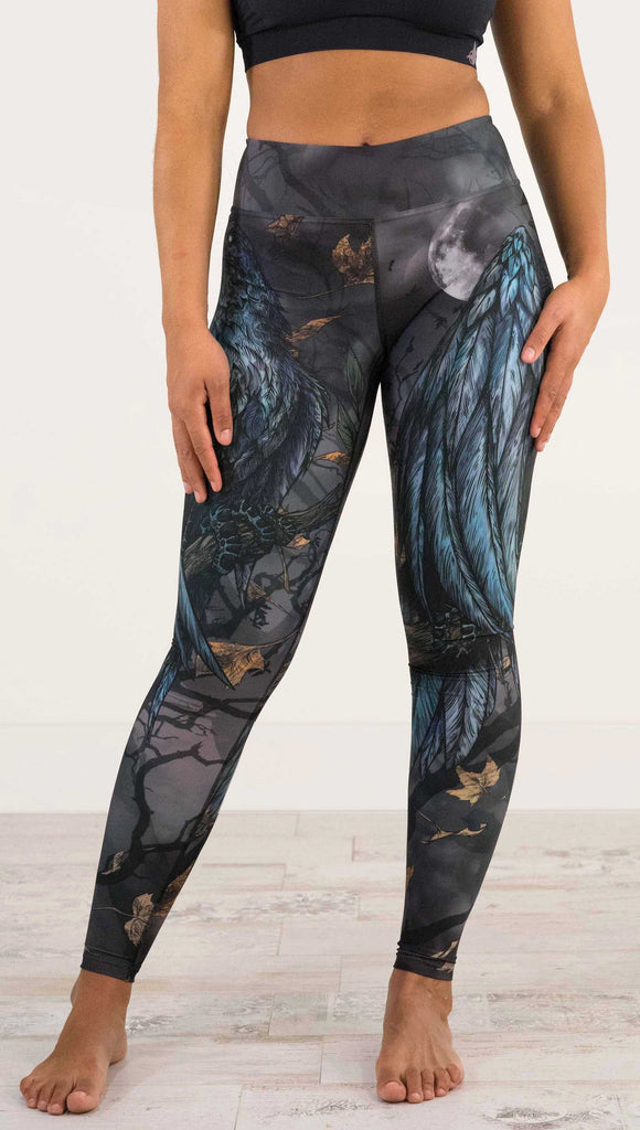 Waist down front view of model wearing WERKSHOP Raven Full Length Triathlon Leggings. The artwork on the leggings is super dark shades of gray and blue with a large raven on the thigh and a dark and stormy moon in the background. Definitely spooky season vibes.
