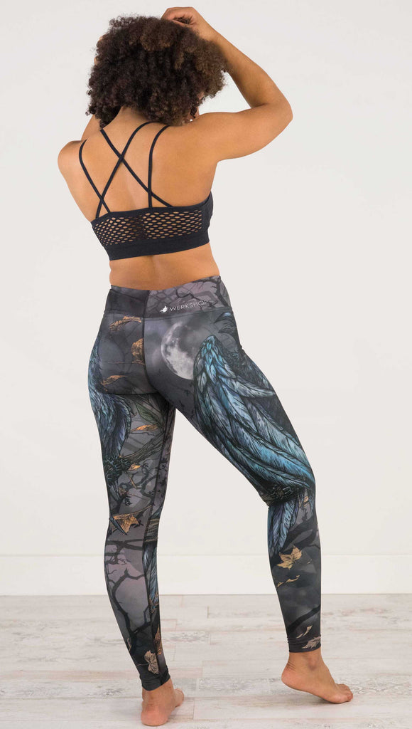 Full body  back view of model wearing WERKSHOP Raven Full Length Triathlon Leggings. The artwork on the leggings is super dark shades of gray and blue with a large raven on the thigh and a dark and stormy moon in the background. Definitely spooky season vibes.