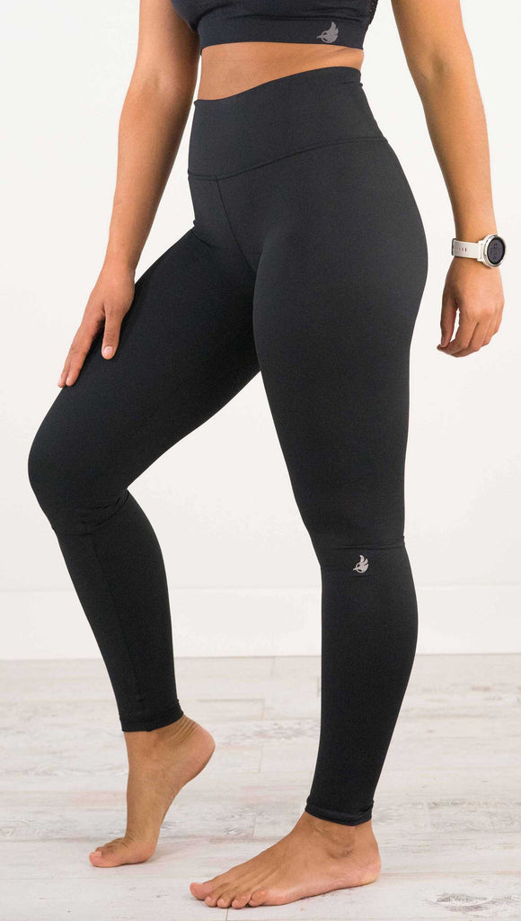 Waist down side view of model wearing WERKSHOP Solid Black Athleisure Leggings with a small reflective eagle logo on the wearers left side calf