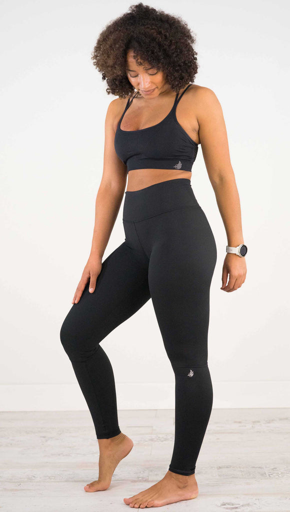 Full body side view of model wearing WERKSHOP Solid Black Athleisure Leggings with a small reflective eagle logo on the wearers left side calf